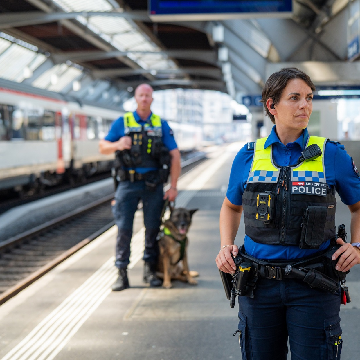Due funzionari delle FFS sono in piedi sulla banchina di una stazione ferroviaria. Uno dei funzionari è più vicino alla telecamera ed è di sesso femminile, quello dietro è di sesso maschile e ha accanto a sé un cane da pastore tedesco